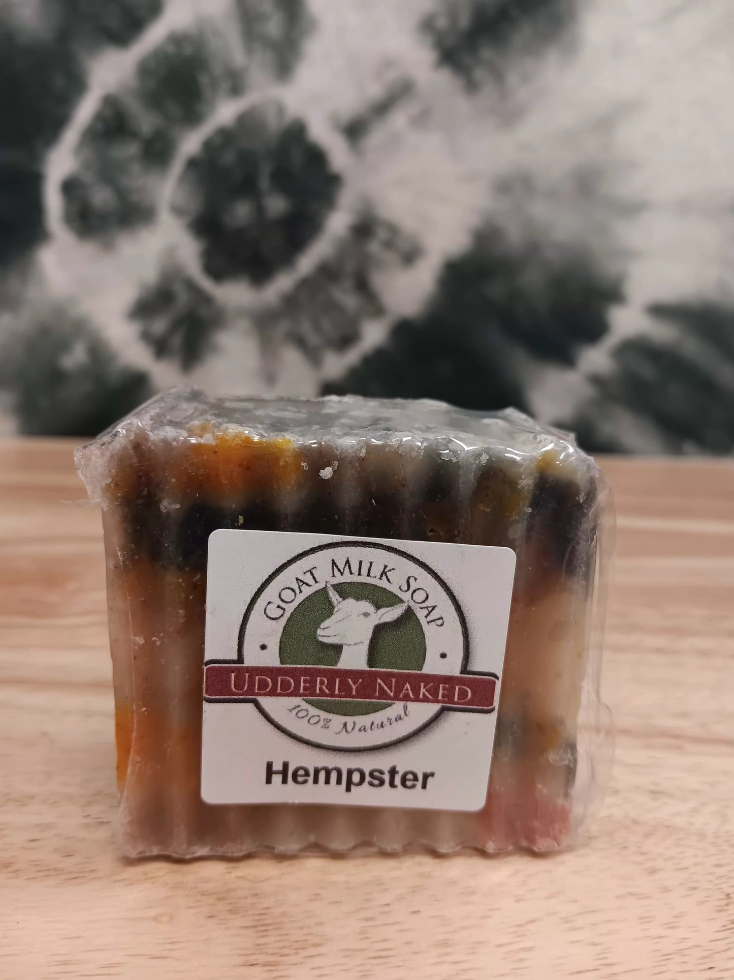 Goat milk soap bar labeled 'Udderly Naked Hempster' on a wooden surface with a blurred natural background.