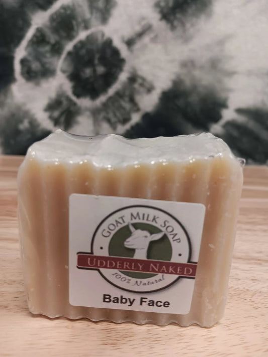 Bar of goat milk soap labeled 'Udderly Naked Baby Face' on a wooden surface with a blurred natural background.